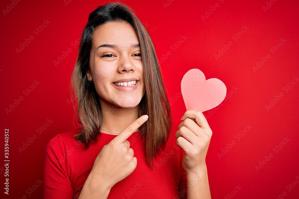 Young beautiful brunette romantic girl holding red paper heart shape over isolated background very happy pointing with hand and finger Young beautiful brunette romantic girl holding red paper heart shape over isolated background very happy pointing with hand and finger