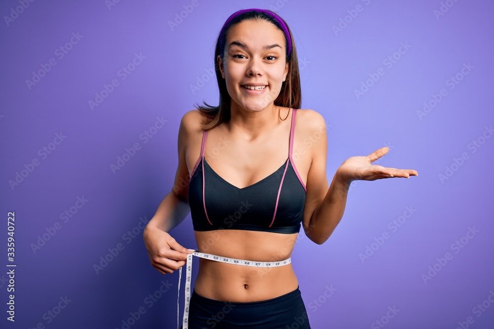 Beautiful slim girl controlling weight using measure tape on waist over purple background very happy and excited, winner expression celebrating victory screaming with big smile and raised hands Beautiful slim girl controlling weight using measure tape on waist over purple background very happy and excited, winner expression celebrating victory screaming with big smile and raised hands