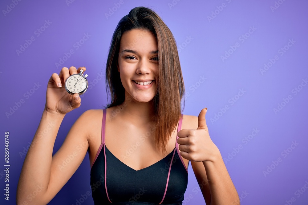Young beautiful sporty girl doing sport using stopwatch over isolated purple background happy with big smile doing ok sign, thumb up with fingers, excellent sign