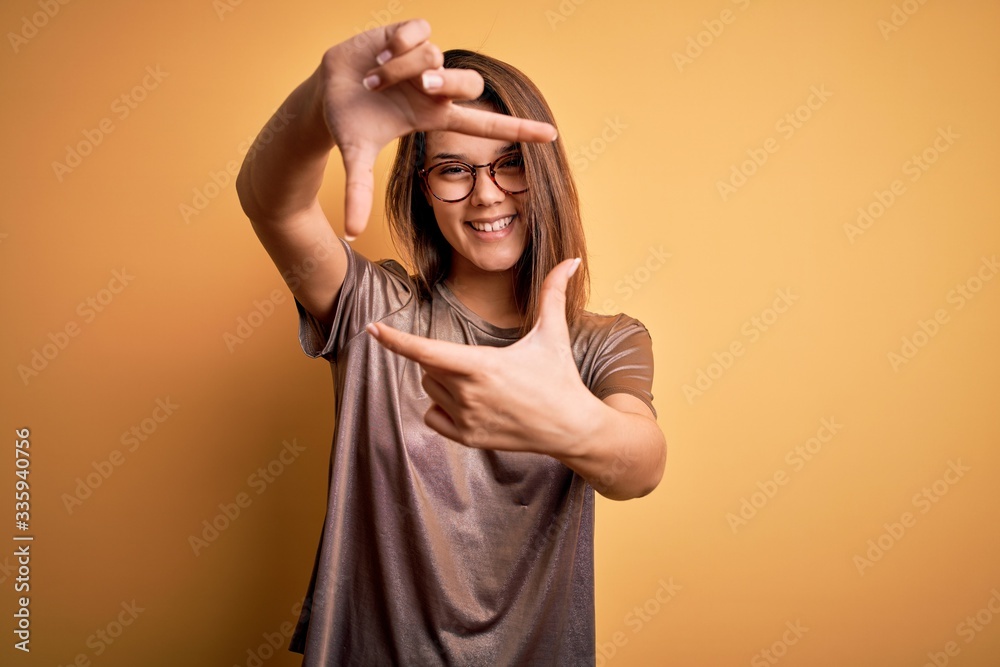 Beautiful brunette girl wearing casual t-shirt and glasses over isolated yellow background smiling making frame with hands and fingers with happy face. Creativity and photography concept. Beautiful brunette girl wearing casual t-shirt and glasses over isolated yellow background smiling making frame with hands and fingers with happy face. Creativity and photography concept.