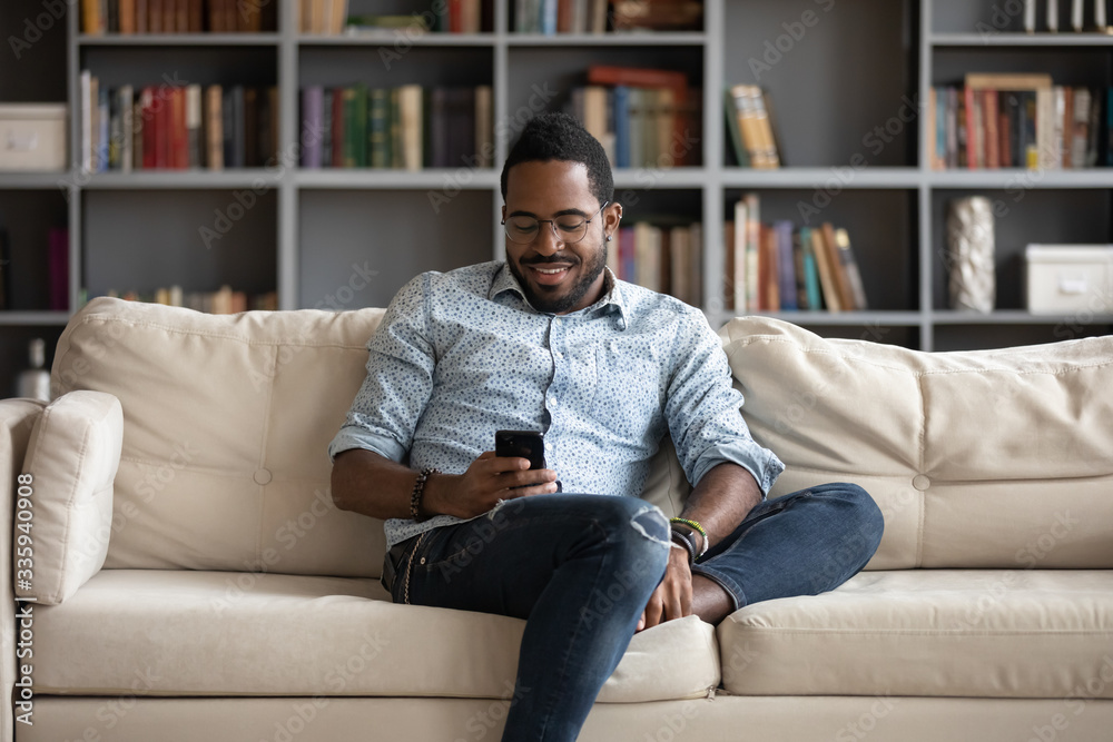 Smiling african American man sit relax on comfortable couch at home using modern cellphone, happy biracial millennial male rest on sofa in living room texting messaging on smartphone gadget - obrazy, fototapety, plakaty 