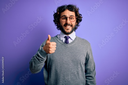 Handsome businessman with beard wearing tie and glasses standing over purple background doing happy thumbs up gesture with hand. Approving expression looking at the camera showing success.
