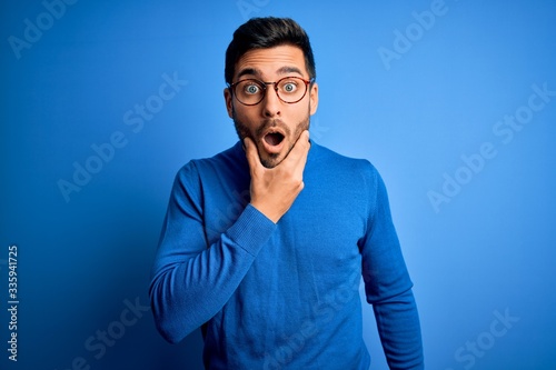 Young handsome man with beard wearing casual sweater and glasses over blue background Looking fascinated with disbelief, surprise and amazed expression with hands on chin