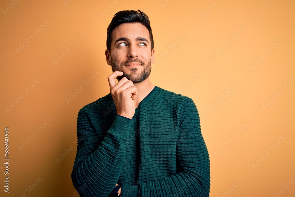 Young handsome man with beard wearing casual sweater standing over yellow background with hand on chin thinking about question, pensive expression. Smiling with thoughtful face. Doubt concept. Young handsome man with beard wearing casual sweater standing over yellow background with hand on chin thinking about question, pensive expression. Smiling with thoughtful face. Doubt concept.