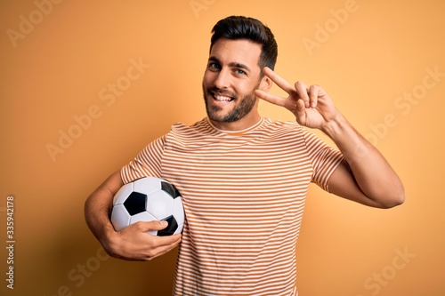 Handsome player man with beard playing soccer holding footballl ball over yellow background Doing peace symbol with fingers over face, smiling cheerful showing victory © Krakenimages.com