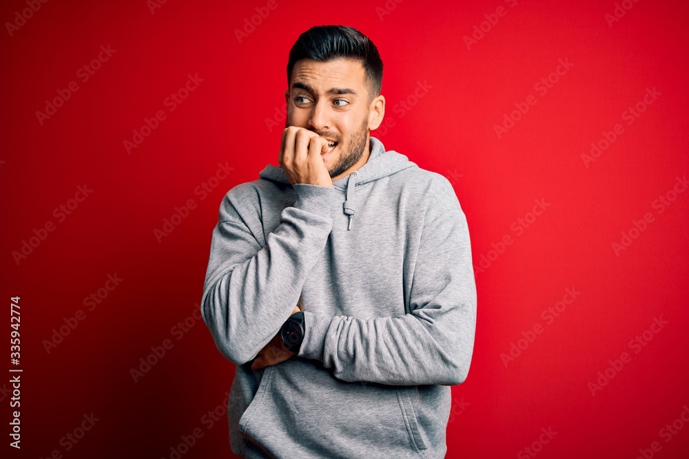 Young handsome sportsman wearing sweatshirt standing over isolated red background looking stressed and nervous with hands on mouth biting nails. Anxiety problem. Young handsome sportsman wearing sweatshirt standing over isolated red background looking stressed and nervous with hands on mouth biting nails. Anxiety problem.