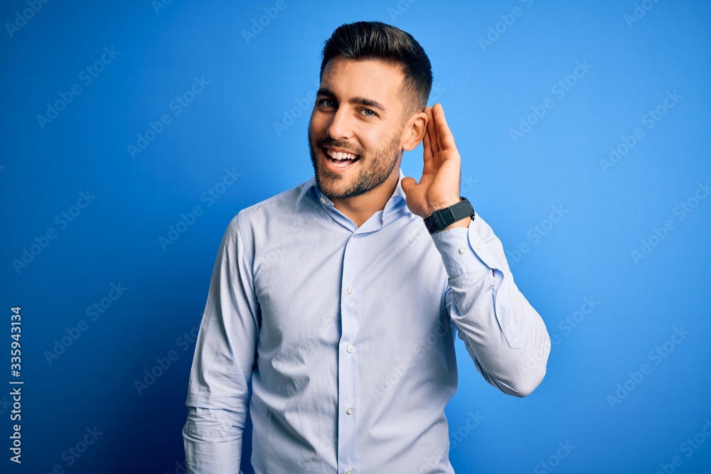 Young handsome man wearing elegant shirt standing over isolated blue background smiling with hand over ear listening an hearing to rumor or gossip. Deafness concept.