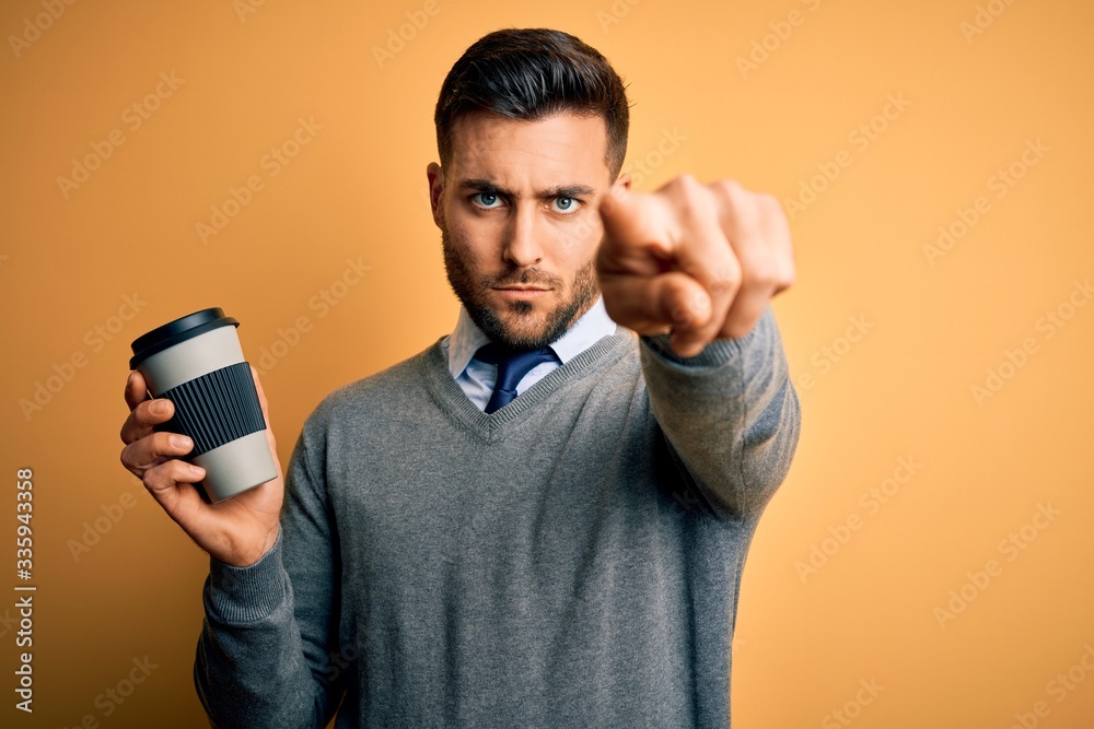 Young handsome man drinking a hot coffee from take away plastic cup over yellow background pointing with finger to the camera and to you, hand sign, positive and confident gesture from the front Young handsome man drinking a hot coffee from take away plastic cup over yellow background pointing with finger to the camera and to you, hand sign, positive and confident gesture from the front