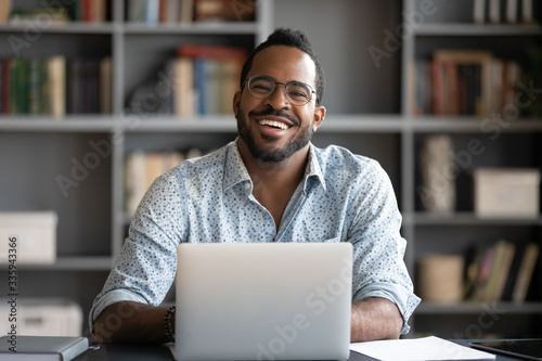 Portrait of smiling African American man in glasses sit at desk in office working on laptop, happy biracial male worker look at camera posing, busy using modern computer gadget at workplace