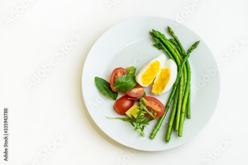 Boiled poached egg, cherry tomatoes, mix salad and asparagus on a white plate on an isolated white background. Healthy Diet Food Concept