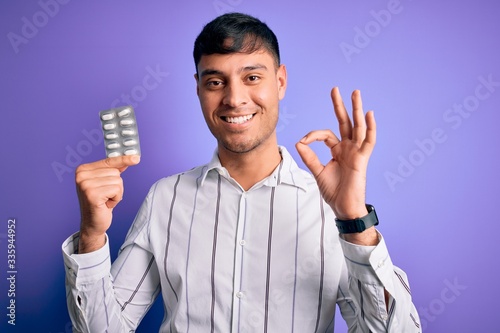 Young hispanic man holding pharmaceutical antibiotics pills over purple isolated background doing ok sign with fingers, excellent symbol