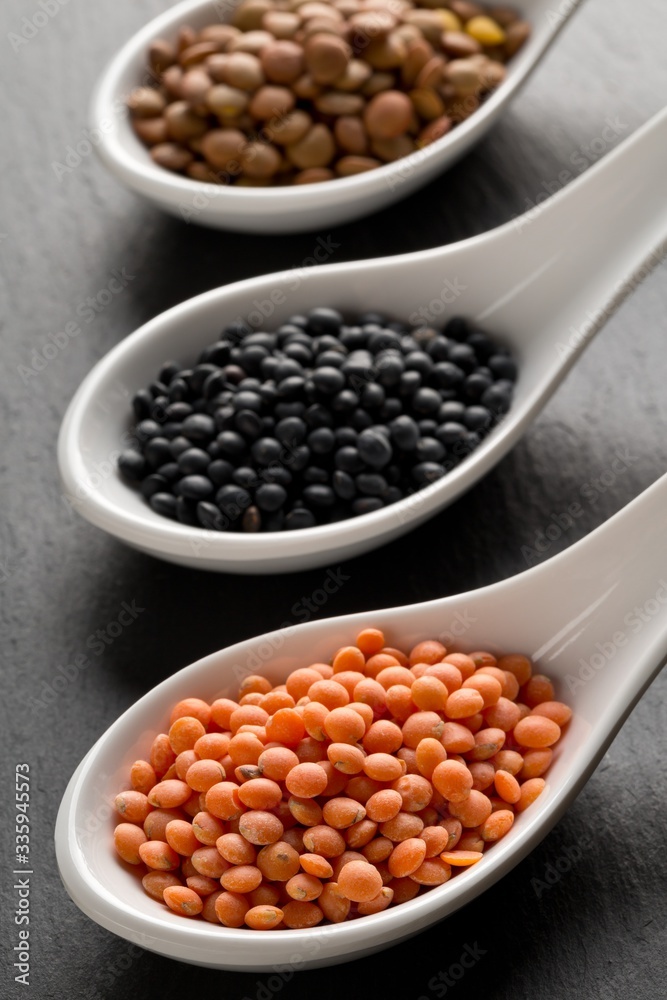 Different assorted lentils mix with red, brown and black beluga lentils in white spoons on dark stone background Different assorted lentils mix with red, brown and black beluga lentils in white spoons on dark stone background