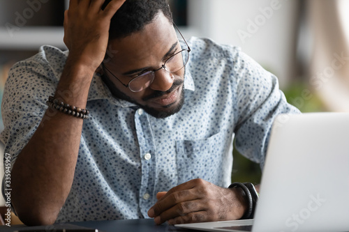 Unhappy tired african American male employee working on laptop look at screen lack motivation or inspiration, unmotivated frustrated biracial man read bad negative news on computer at workplace