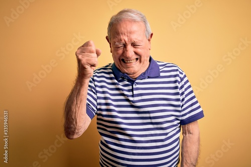 Grey haired senior man wearing casual navy striped t-shirt standing over yellow background angry and mad raising fist frustrated and furious while shouting with anger. Rage and aggressive concept.