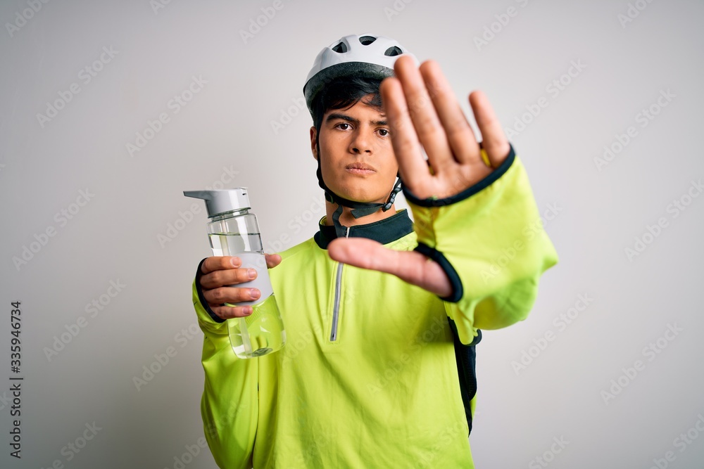 Young handsome cyclist man wearing security bike helmet drinking bottle of water with open hand doing stop sign with serious and confident expression, defense gesture Young handsome cyclist man wearing security bike helmet drinking bottle of water with open hand doing stop sign with serious and confident expression, defense gesture