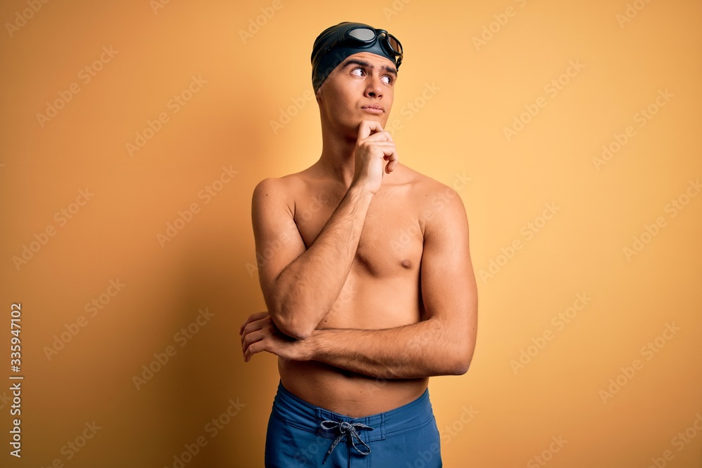 Young handsome man shirtless wearing swimsuit and swim cap over isolated yellow background with hand on chin thinking about question, pensive expression. Smiling with thoughtful face. Doubt concept.