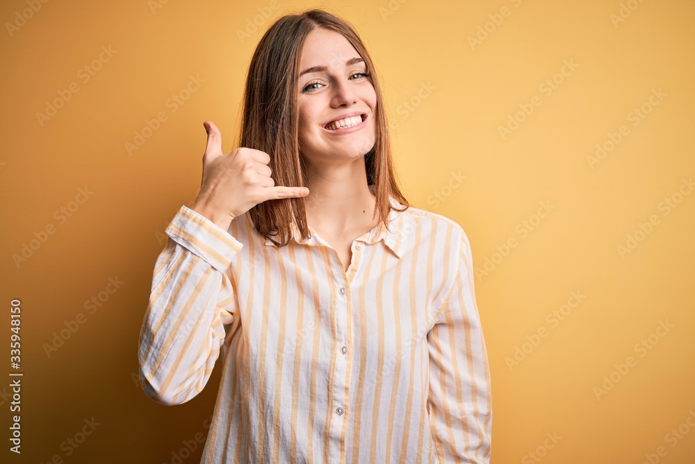 Young beautiful redhead woman wearing casual striped shirt over isolated yellow background smiling doing phone gesture with hand and fingers like talking on the telephone. Communicating concepts. Young beautiful redhead woman wearing casual striped shirt over isolated yellow background smiling doing phone gesture with hand and fingers like talking on the telephone. Communicating concepts.