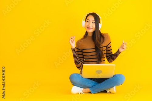 Beautiful portrait young asian woman sit on the floor with laptop and headphone on yellow background © siraphol
