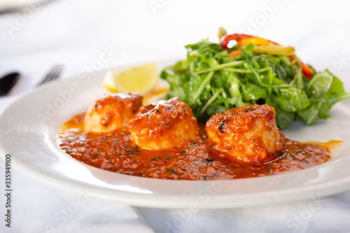 A view of a plate of large scallops in Arrabbiata sauce, next to an arugula salad, in a restaurant or kitchen setting.