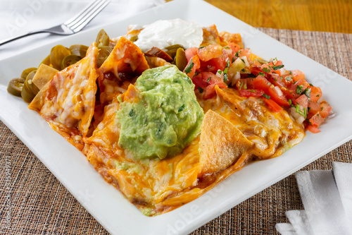 A view of a plate of loaded nachos, with a big dollop of guacamole, in a restaurant or kitchen setting.