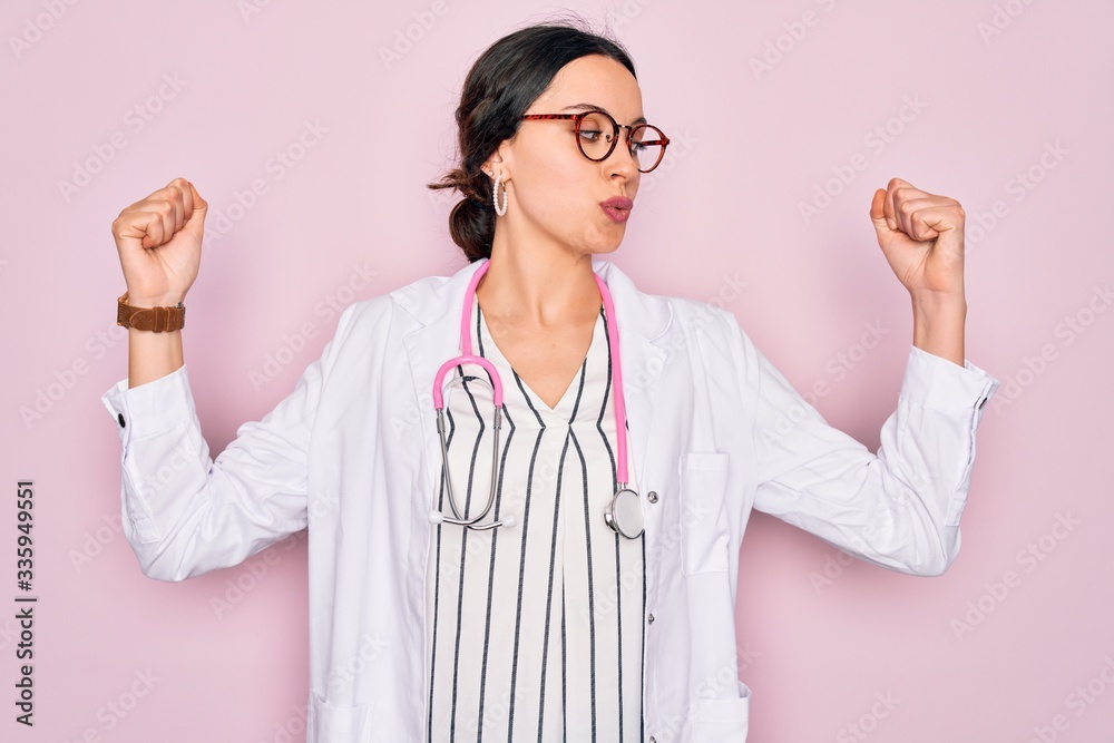 Beautiful doctor woman with blue eyes wearing coat and stethoscope over pink background showing arms muscles smiling proud. Fitness concept. Beautiful doctor woman with blue eyes wearing coat and stethoscope over pink background showing arms muscles smiling proud. Fitness concept.