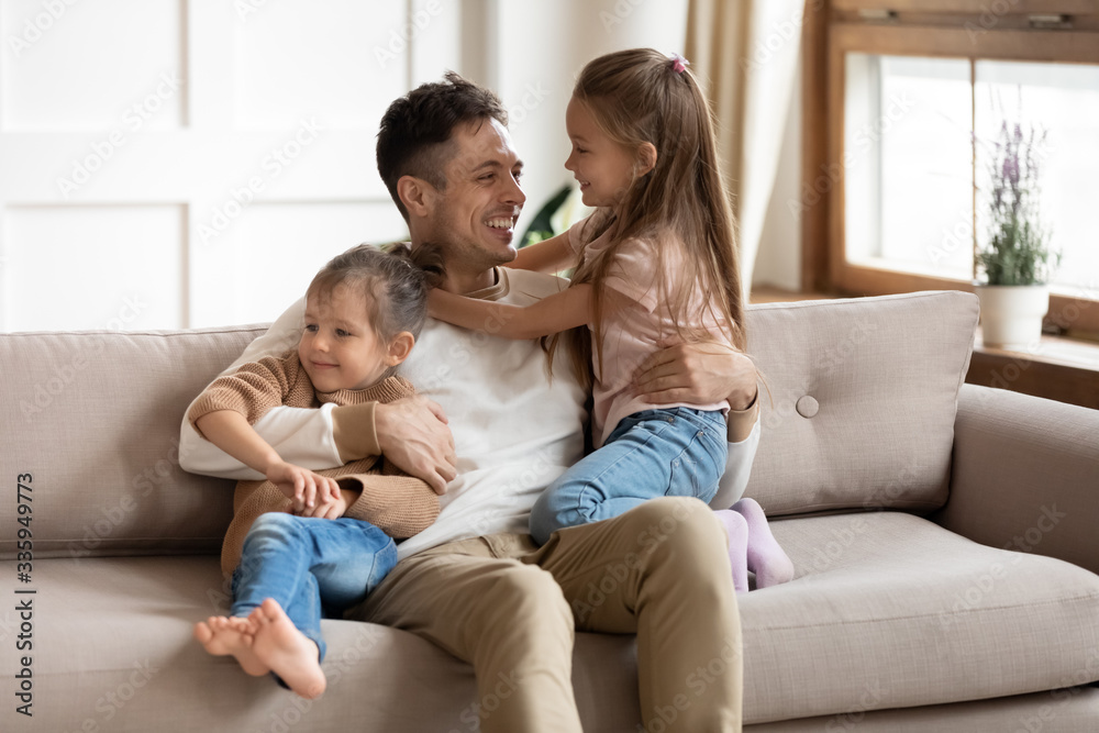 Smiling young father sitting on comfortable couch, playing with small cute kids daughters in living room. Happy uncle having fun communicating entertaining with little adorable children nephews. Smiling young father sitting on comfortable couch, playing with small cute kids daughters in living room. Happy uncle having fun communicating entertaining with little adorable children nephews.