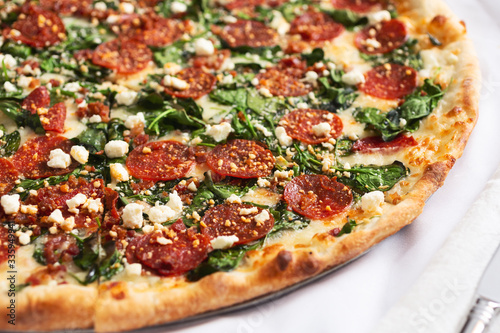 A closeup view of a Greek style pizza pie, featuring pepperoni, spinach and feta cheese, in a restaurant or kitchen setting.
