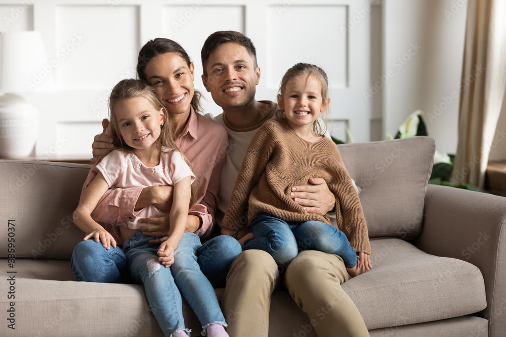 Happy young couple holding on lap cute small kids daughters, looking at camera. Portrait of adorable family of four relaxing on comfortable sofa, posing for photo together in living room at home.