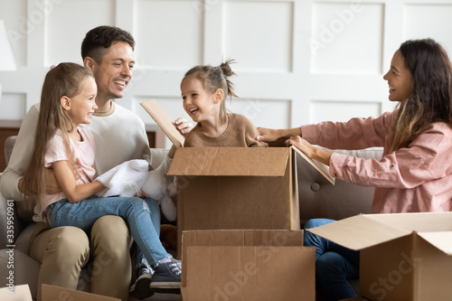 Adorable playful little girl hiding in big cardboard box, while happy parents and sister finding her. Joyful family couple having fun with cute children in living room after moving in new apartment.