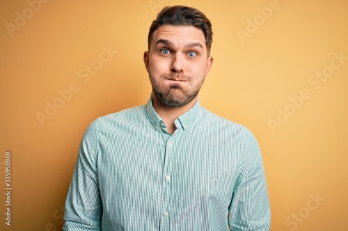 Young business man with blue eyes wearing elegant green shirt over yellow background puffing cheeks with funny face. Mouth inflated with air, crazy expression.
