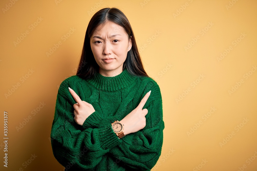 Young beautiful asian woman wearing green winter sweater over yellow isolated background Pointing to both sides with fingers, different direction disagree Young beautiful asian woman wearing green winter sweater over yellow isolated background Pointing to both sides with fingers, different direction disagree
