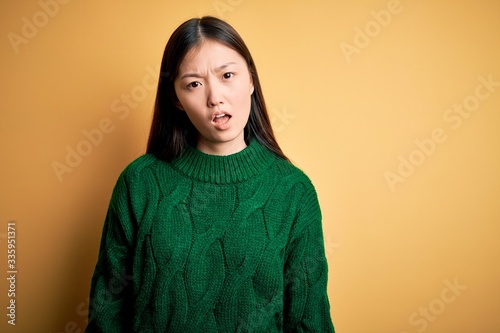 Young beautiful asian woman wearing green winter sweater over yellow isolated background In shock face, looking skeptical and sarcastic, surprised with open mouth © Krakenimages.com
