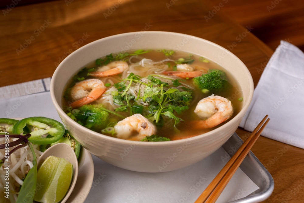 A view of a pho soup bowl with shrimp and vegetables, in a restaurant or kitchen setting.