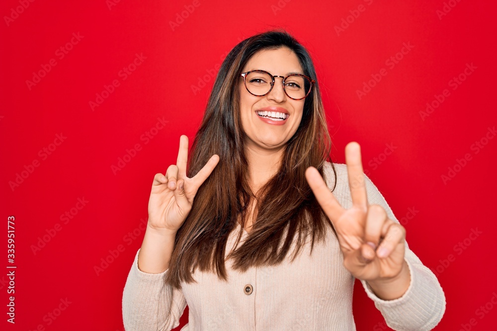 Young hispanic smart woman wearing glasses standing over red isolated background smiling looking to the camera showing fingers doing victory sign. Number two. Young hispanic smart woman wearing glasses standing over red isolated background smiling looking to the camera showing fingers doing victory sign. Number two.