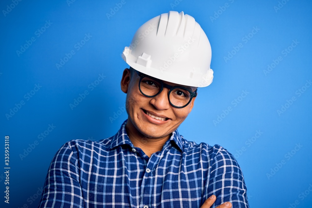 Young handsome engineer latin man wearing safety helmet over isolated blue background happy face smiling with crossed arms looking at the camera. Positive person. Young handsome engineer latin man wearing safety helmet over isolated blue background happy face smiling with crossed arms looking at the camera. Positive person.