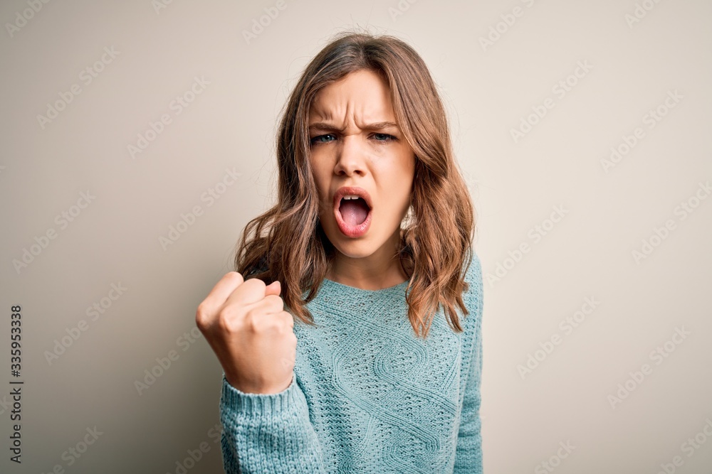 Young blonde girl wearing casual blue winter sweater over isolated background angry and mad raising fist frustrated and furious while shouting with anger. Rage and aggressive concept. Young blonde girl wearing casual blue winter sweater over isolated background angry and mad raising fist frustrated and furious while shouting with anger. Rage and aggressive concept.