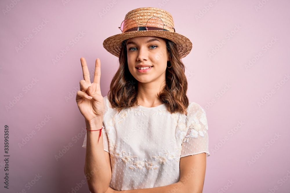 Young beautiful blonde girl wearing summer hat over pink isolated background smiling with happy face winking at the camera doing victory sign. Number two. Young beautiful blonde girl wearing summer hat over pink isolated background smiling with happy face winking at the camera doing victory sign. Number two.