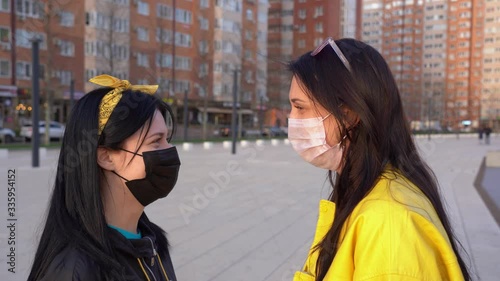 Two cute women wear medical protective face masks look in love each other with care and talk on city background at sunset close up. Girl in black jacket straighten pink mask her brunette girlfriend. photo