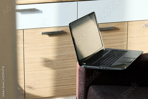 An open black laptop stands on the armrest of a sofa against a blurred background of furniture in the room during the day