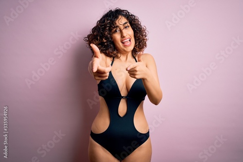 Young beautiful arab woman on vacation wearing swimsuit and sunglasses over pink background pointing fingers to camera with happy and funny face. Good energy and vibes.