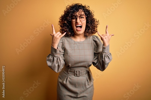 Beautiful arab business woman wearing dress and glasses standing over yellow background shouting with crazy expression doing rock symbol with hands up. Music star. Heavy music concept. © Krakenimages.com
