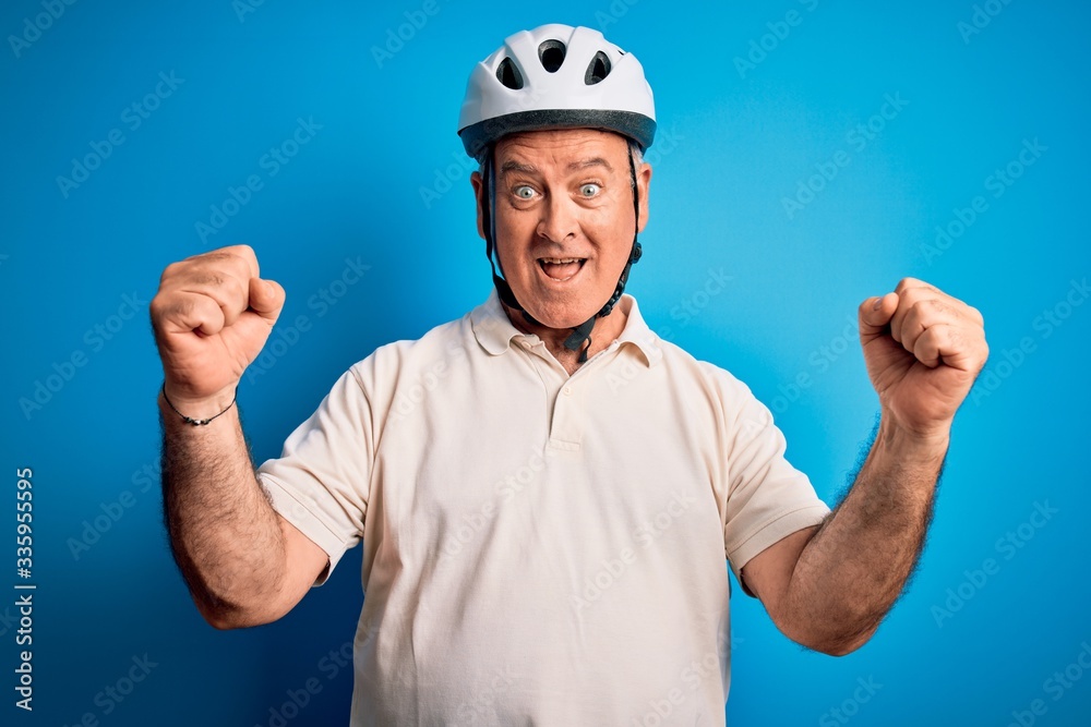 Middle age hoary cyclist man wearing bike security helmet over isolated blue background celebrating surprised and amazed for success with arms raised and open eyes. Winner concept. Middle age hoary cyclist man wearing bike security helmet over isolated blue background celebrating surprised and amazed for success with arms raised and open eyes. Winner concept.