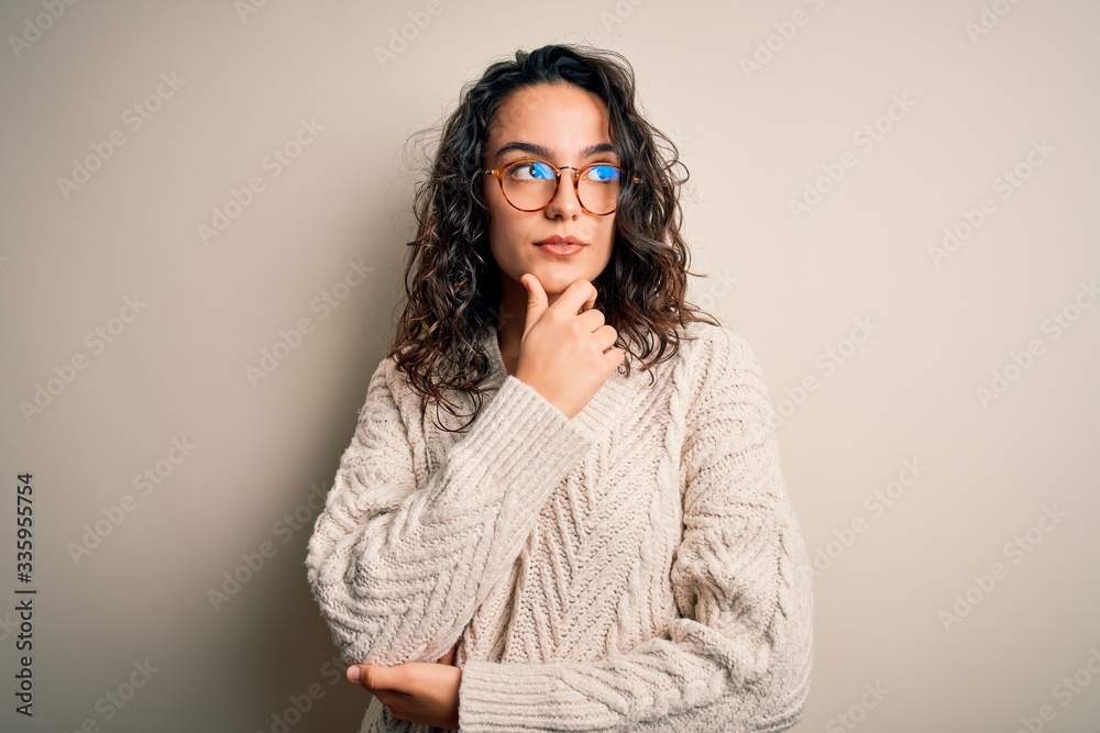 Beautiful woman with curly hair wearing casual sweater and glasses over white background with hand on chin thinking about question, pensive expression. Smiling with thoughtful face. Doubt concept.