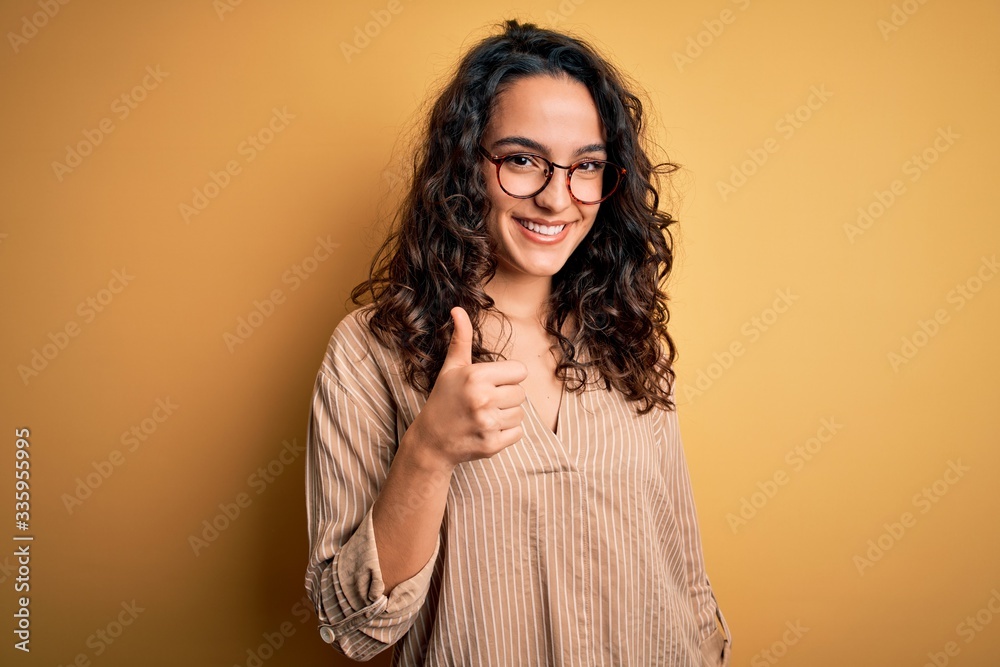 Beautiful woman with curly hair wearing striped shirt and glasses over yellow background doing happy thumbs up gesture with hand. Approving expression looking at the camera showing success. Beautiful woman with curly hair wearing striped shirt and glasses over yellow background doing happy thumbs up gesture with hand. Approving expression looking at the camera showing success.