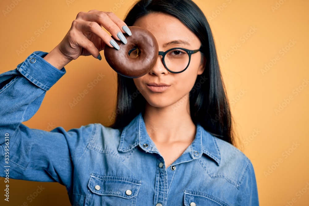 Young beautiful chinese woman holding chocolate donut over isolated yellow background with a confident expression on smart face thinking serious Young beautiful chinese woman holding chocolate donut over isolated yellow background with a confident expression on smart face thinking serious