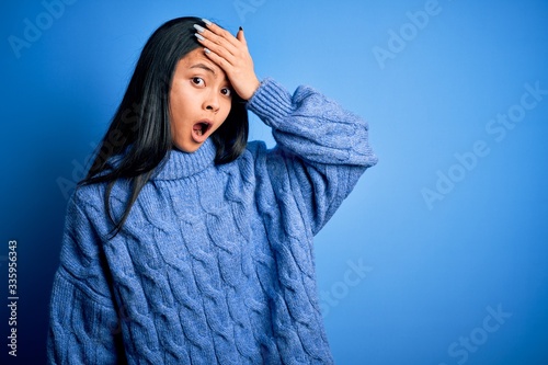 Young beautiful chinese woman wearing casual sweater over isolated blue background surprised with hand on head for mistake, remember error. Forgot, bad memory concept.