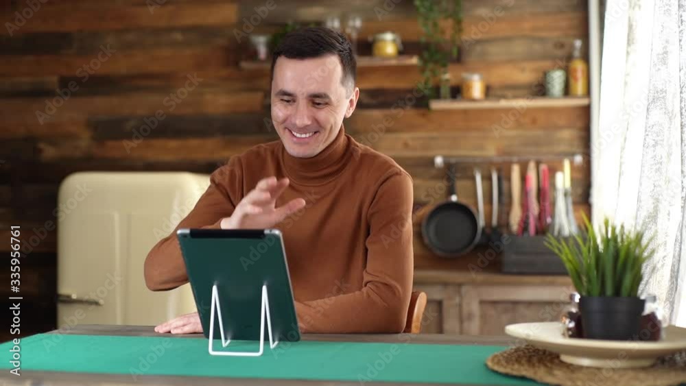 Handsome Caucasian young man using tablet computer having online video chat with friends while sitting at the table in cozy room. Shooting in slow motion. Handsome Caucasian young man using tablet computer having online video chat with friends while sitting at the table in cozy room. Shooting in slow motion.