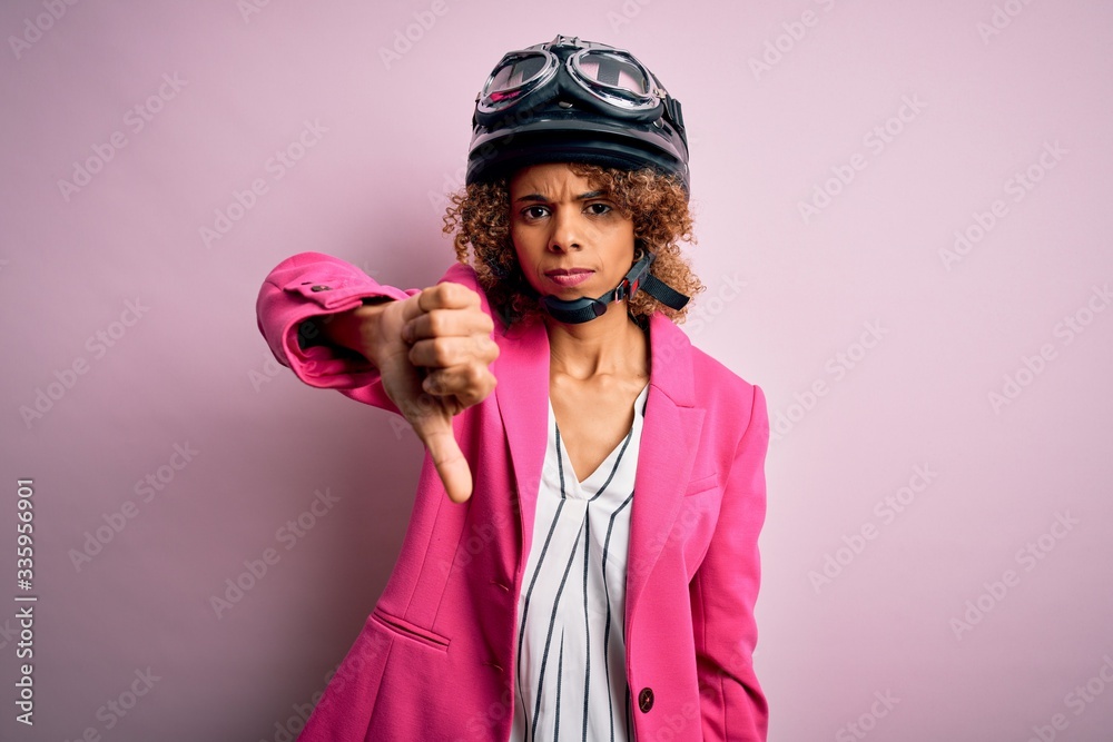 African american motorcyclist woman with curly hair wearing moto helmet over pink background looking unhappy and angry showing rejection and negative with thumbs down gesture. Bad expression. African american motorcyclist woman with curly hair wearing moto helmet over pink background looking unhappy and angry showing rejection and negative with thumbs down gesture. Bad expression.
