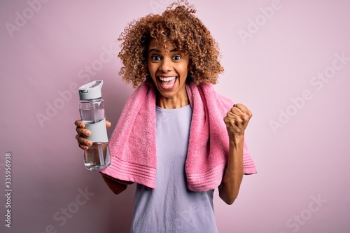 Young african american sportswoman doing sport wearing towel drinking bottle of water screaming proud and celebrating victory and success very excited, cheering emotion