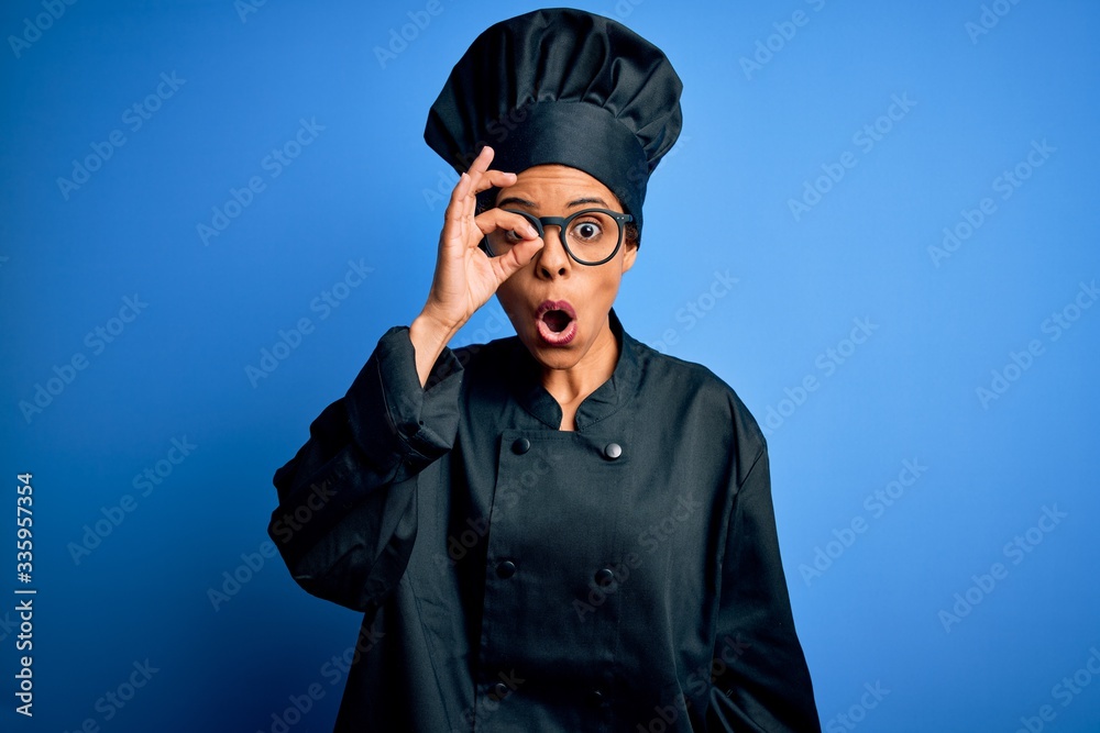 Young african american chef woman wearing cooker uniform and hat over blue background doing ok gesture shocked with surprised face, eye looking through fingers. Unbelieving expression. Young african american chef woman wearing cooker uniform and hat over blue background doing ok gesture shocked with surprised face, eye looking through fingers. Unbelieving expression.